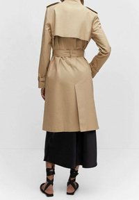 Cappotto beige con vita abbottonata, spalline e una pattina sul retro; abbinato a una gonna nera in satin e sandali con cinturini.