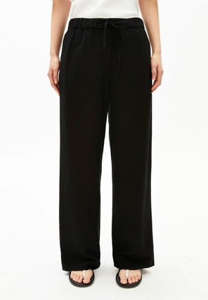 Persoon die losse zwarte broek met trekkoord en zwarte slippers draagt, staand tegen een witte achtergrond.