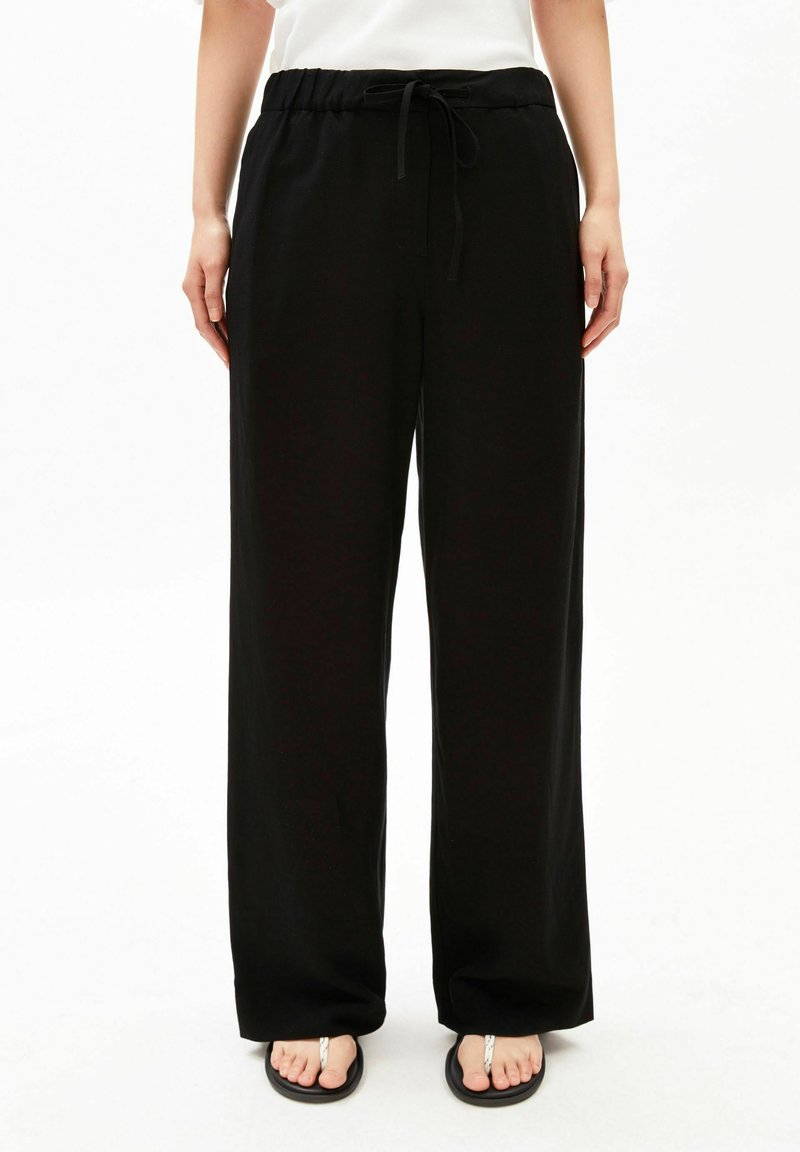 Person wearing loose black drawstring pants and black flip-flops standing against a white background.