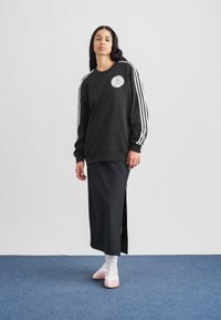 Μαύρο φούτερ με λευκές ρίγες, κυκλικό γραφικό στο στήθος, συνδυασμένο με μακριά μαύρη φούστα με πλευρικό σκίσιμο και ανοιχτό ροζ παπούτσια.