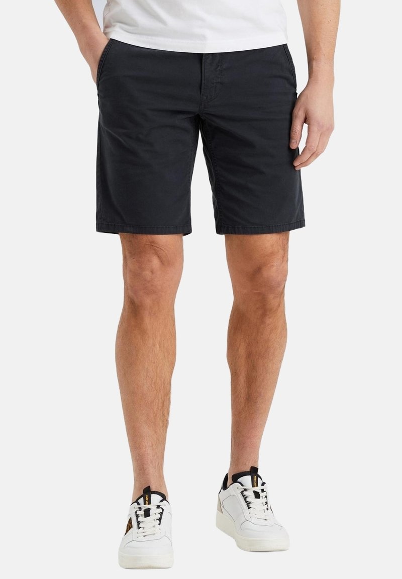 Homme portant un short noir jusqu'aux genoux, les mains dans les poches, et des baskets blanches avec des accents noirs sur fond blanc.