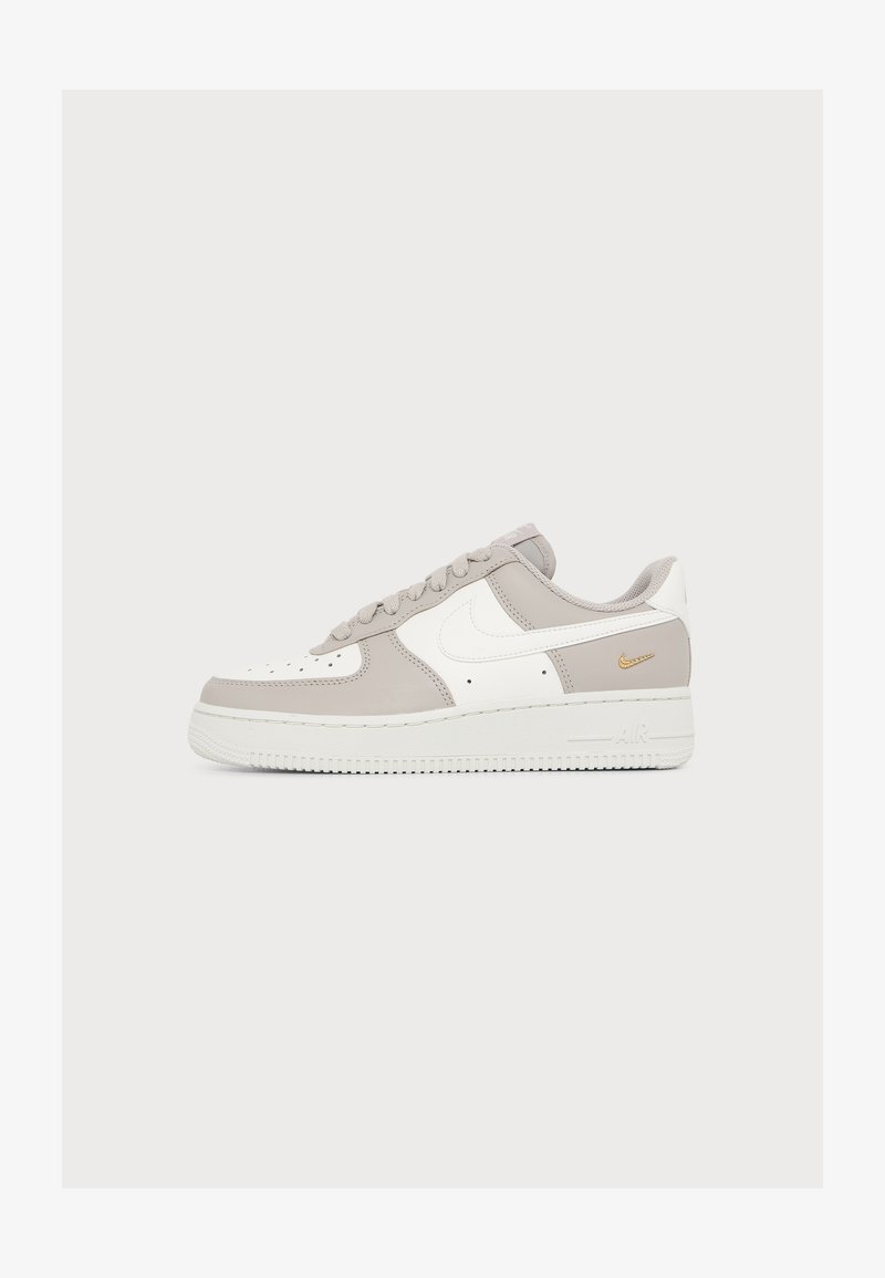 Χαμηλό sneaker Nike Air Force 1 σε λευκό και ανοιχτό μπεζ με λευκό swoosh και μικρό χρυσό λογότυπο κοντά στη φτέρνα, εμφανίζεται από την εξωτερική πλευρά.