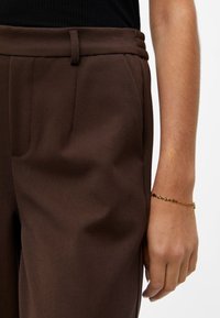 Un pantalon marron sur mesure présente une texture lisse, des poches latérales et une ceinture avec des détails cousus. Un bracelet floral doré orne le poignet.