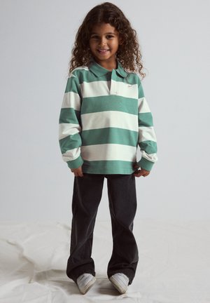 Jong kind met krullend haar, gekleed in een groen-wit gestreept shirt met lange mouwen, zwarte broek en witte sneakers, glimlachend naar de camera.