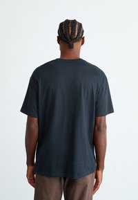 Homme avec des cheveux tressés portant un t-shirt noir uni et un pantalon marron, se tenant face à un fond blanc, dos au spectateur.