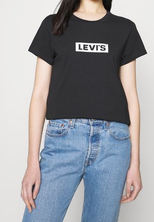 Vrouw draagt een zwart Levi's T-shirt en lichtblauwe high-waisted jeans tegen een effen lichte achtergrond.