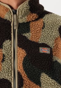 Närbild av en fleecejacka med brunt, grönt och tan kamouflage mönster och en framdragkedja, prydd med en liten Dickies-logotypspatch.