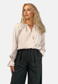 Jasnoróżowa bluzka z dekoltem w kształcie litery V, zebranymi ramionami i długimi rękawami, w połączeniu z ciemnymi spodniami w prążki z zakładkami. Akcent w postaci złotego łańcuszka.