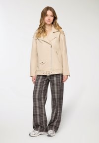 Giacca biker in pelle beige chiaro con ferramenta argentata, tasche con zip e vita con cintura, abbinata a pantaloni a gamba larga grigi a quadri.