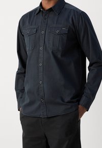 Dunkelblaues Denim-Hemd mit einem Button-Down-Kragen, zwei Brusttaschen und kontrastierenden Knöpfen. Lange Ärmel mit geknöpften Manschetten.