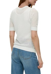 Lekki, biały sweter dzianinowy z falistym wzorem, krótkie rękawy oraz ściągacze na dekolcie i u dołu. Połączony z niebieskimi dżinsami.