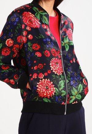 Personne portant une veste bomber bleu marine avec des motifs floraux rouges, verts et violets et une chemise rouge en dessous.