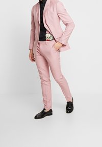 Costume rose clair avec une coupe ajustée, comprenant un blazer croisé, un pantalon à plat devant, et une chemise noire avec des accents floraux à l'intérieur. Mocassins noirs.