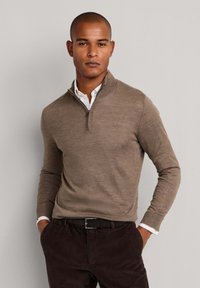 Bruine, met rits gesloten pullover trui met een gestructureerd breisel, voorzien van een hoge kraag en een gelaagde witte blouse eronder, gecombineerd met donkere broek.