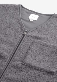 Veste zippée grise avec un extérieur en polaire texturé, un col en V et une poche à patch sur le côté gauche. Présente des fermetures éclair en métal.