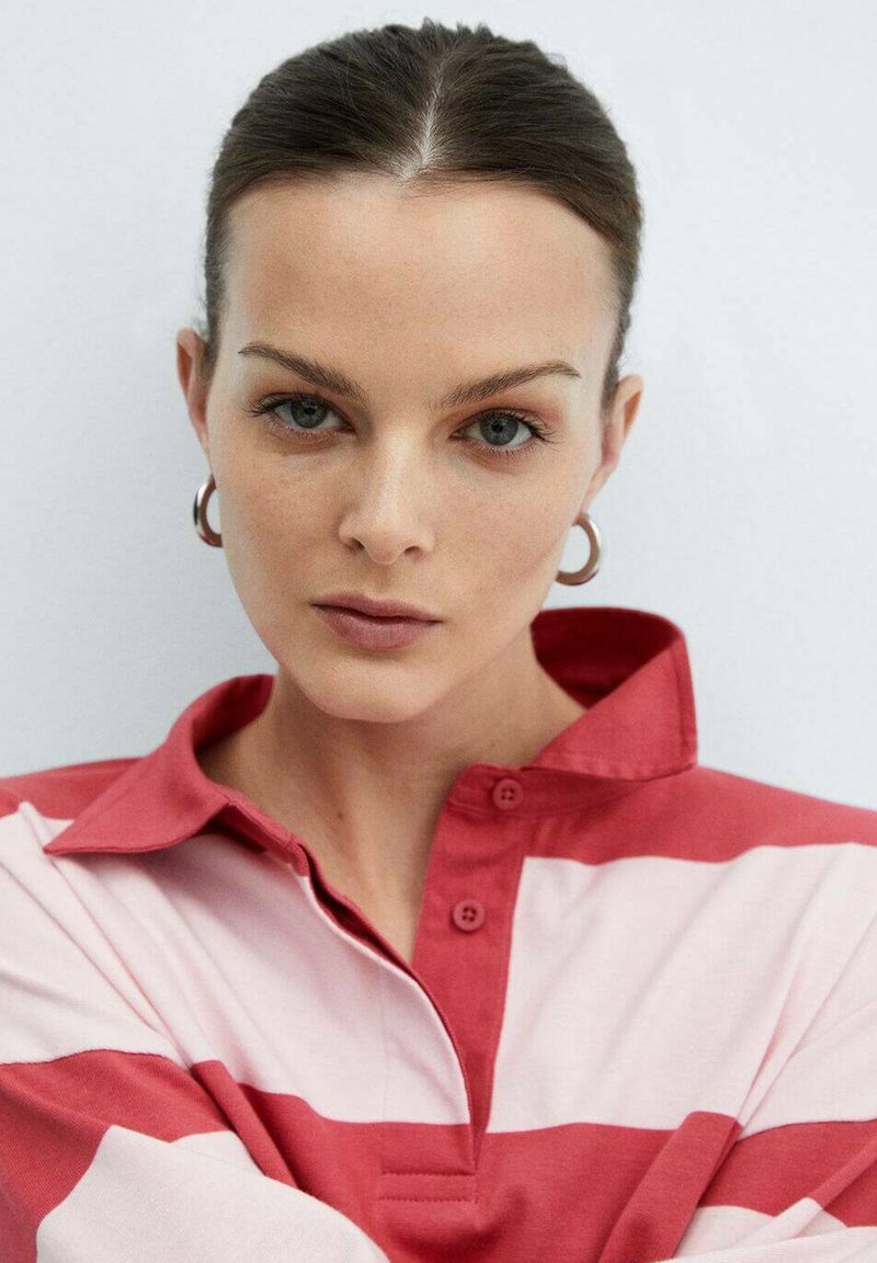 Camicia a righe rosse e bianche con colletto e chiusura a bottoni, realizzata in tessuto morbido. Il modello ha i capelli di lunghezza media e indossa orecchini a cerchio.