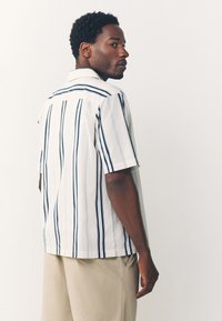 Chemise à manches courtes blanche avec des rayures verticales bleu marine et bleu clair, dotée d'un col pointu et d'une coupe décontractée.