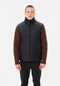 Schwarze ärmellose Steppweste mit hohem Kragen, gestepptem Design und Frontreißverschluss, kombiniert mit einem braunen Langarmshirt darunter.