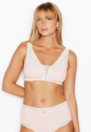 Donna con capelli biondi che indossa un reggiseno rosa chiaro con rifiniture in pizzo e mutandine a vita alta abbinate, in piedi davanti a uno sfondo bianco.