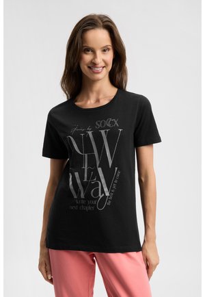 Lächelnde Frau mit braunen Haaren, die ein schwarzes T-Shirt mit Grafik und pinke Hosen trägt, vor einem einfarbigen hellen Hintergrund stehend.