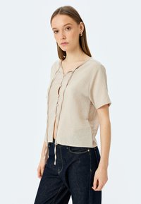 Camisa de lino beige de manga corta con cuello redondo y botonadura frontal, dotada de lazos en el dobladillo. Acompañada de jeans de mezclilla oscuros.