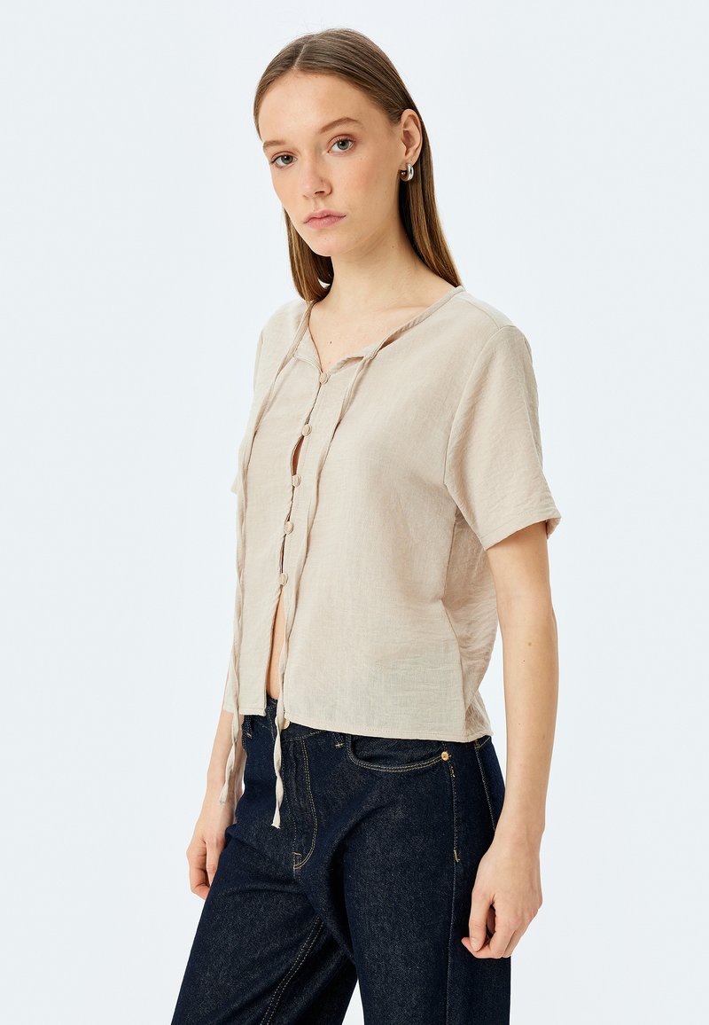 Camisa de lino beige de manga corta con cuello redondo y botonadura frontal, dotada de lazos en el dobladillo. Acompañada de jeans de mezclilla oscuros.