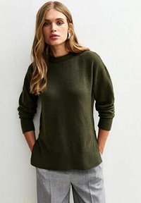Maglione lavorato a maglia di un verde scuro con scollatura rotonda e maniche lunghe, abbinato a pantaloni grigi chiari. Texture morbida e vestibilità rilassata.