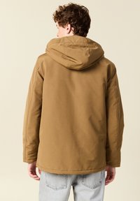 Joven con cabello rizado que lleva una chaqueta con capucha marrón y jeans de lavado claro, de espaldas contra un fondo beige liso.