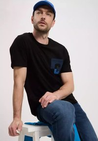 Czarny t-shirt z krótkim rękawem z niebieską kieszenią z literą C. Ubrany w niebieskie dżinsy i niebieską czapkę, siedzący na białym stołku.