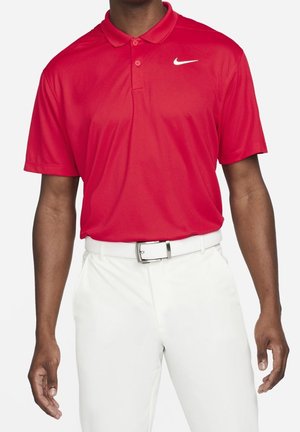 Mężczyzna w czerwonej koszulce polo Nike z białym logo Swoosh, włożonej w białe spodnie z białym paskiem i srebrną klamrą.