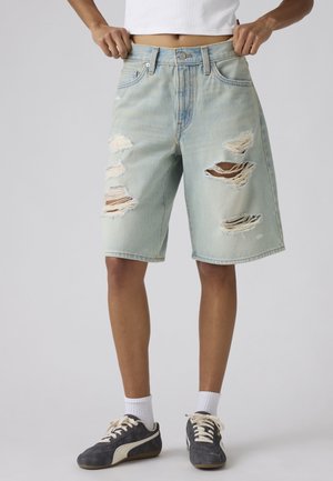 Personne portant un short en jean bleu clair déchiré, des chaussettes blanches, des baskets noires et un top court blanc, debout devant un fond uni.