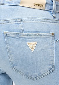 Πίσω τσέπη από ανοιχτό μπλε τζιν Guess που δείχνει το τριγωνικό σήμα Guess και μπεζ δερμάτινη ετικέτα με την επωνυμία στη μέση.