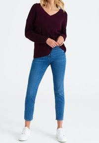 Nainen, jolla on päällään tummanvioletti V-pääntie neule, siniset slim fit farkut ja valkoiset tennarit, seisoo kädet vyötärön lähellä valkoista taustaa vasten.