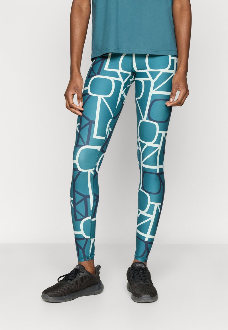 Στενά κολάν σε τυρκουάζ με μεγάλα λευκά γεωμετρικά σχέδια. Κατασκευασμένα από ελαστικό υλικό, συνδυασμένα με μαύρα αθλητικά παπούτσια.