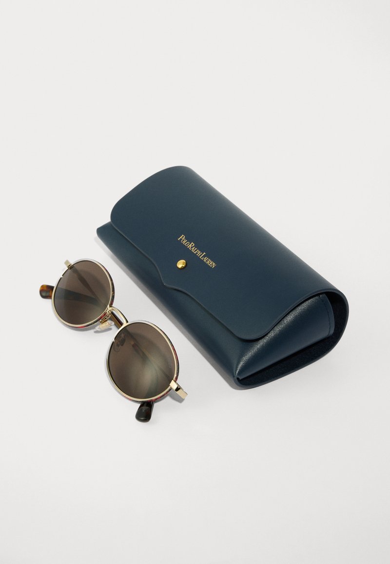 Sunglasses with round dark lenses, gold metal frame, and tortoiseshell accents, beside a dark blue leather case with a gold logo.