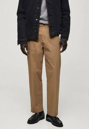 Beige Hosen in einer lockeren Passform, kombiniert mit einem hellgrauen Sweatshirt und einer schwarzen Denimjacke. Schwarze Slipper runden das Outfit ab.