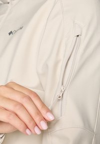 Veste beige clair en tissu doux, dotée d'une poche zippée sur la manche et d'une texture lisse, avec des détails de couture minimaux.