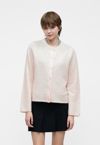 Νεαρή γυναίκα ντυμένη με ένα ανοιχτό ροζ πουκάμισο cardigan στερεωμένο με κουμπιά πάνω από ένα μπλουζάκι και μια μαύρη πλισέ φούστα, στέκεται μπροστά σε ένα απλό λευκό φόντο.