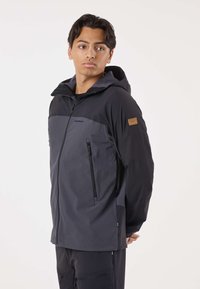 Giovane uomo che indossa una giacca con cappuccio a due toni, grigio scuro e nero, con tasche con cerniera, che guarda di lato su uno sfondo semplice.