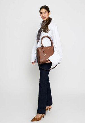 Woman wearing white blouse and dark jeans, holding a brown leather handbag with top handles and shoulder strap, standing against a plain background.