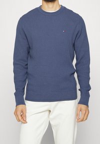 Maglione blu a coste con maniche lunghe e colletto a girocollo a coste. Presenta un piccolo logo sul petto. Indossato su pantaloni chiari.