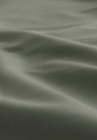 Hladká, zelená tkanina s mírným leskem a jemnou texturou, vykazující jemné vlny a záhyby, což naznačuje flexibilitu a vzhled podobný kůži.