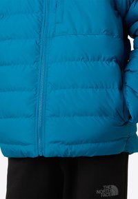 Kék pufi dzseki vízszintes varrással, cipzáras záródással és oldalzsebekkel. Fekete nadrággal párosítva, amely logóval van ellátva. Simább textúra.