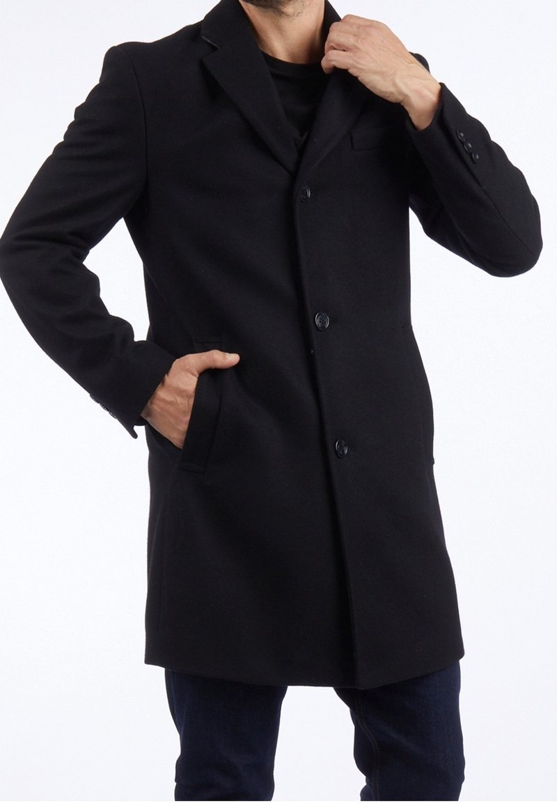 Cappotto lungo in misto lana nero con revers a cascata, chiusura con bottoni e tasche laterali. Il design aderente e la texture liscia creano un aspetto sartoriale.