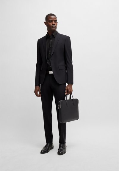 Costume noir à coupe ajustée, associé à une chemise noire. Porte un sac en cuir noir rectangulaire, avec des chaussures noires brillantes pour un look formel.