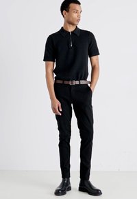 Črna polo majica s kratkimi rokavi in zadrgo na ovratniku, v kombinaciji s črnimi slim-fit hlačami in črnimi usnjenimi gležnjarji.