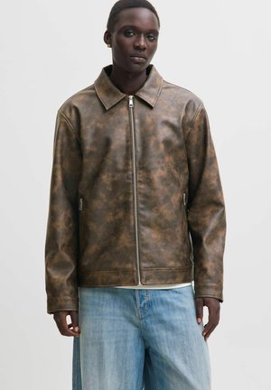 Personne portant une veste en cuir marron usée avec une fermeture éclair à l'avant et un jean bleu, debout devant un fond clair uni.