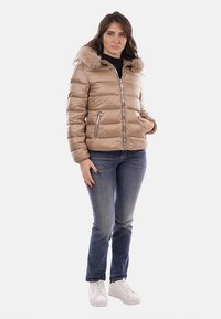 Giacca imbottita marrone con cappuccio foderato in pelliccia sintetica, chiusura frontale con cerniera e due tasche laterali. Abbinata a jeans blu e scarpe da ginnastica bianche.