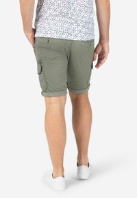 Olive green cargo shorts with rolled hems, multiple pockets, and a textured fabric. Worn with white sneakers and a patterned white shirt.