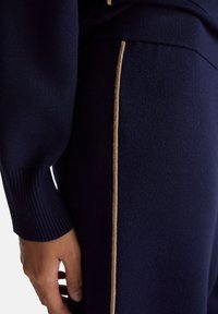 Granatowe dzianinowe spodnie o prążkowanej fakturze, z kontrastowym złoto-beżowym paskiem bocznym wzdłuż szwu, podkreślającym design.